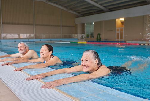 Group of active seniors holding poolside splashing feet exercising in indoor pool