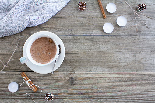 Hot chocolate stirred with a cinnamon stick with candles, cinnamon and a cozy woollen jumper.\nShot from above on a rustic wooden table with copy space. Flat lay.