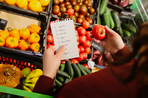woman chooses vegetables in supermarket with list of products to buy. Bright fruit counter on background. leaf from notebook and tomato in hands. Top view.