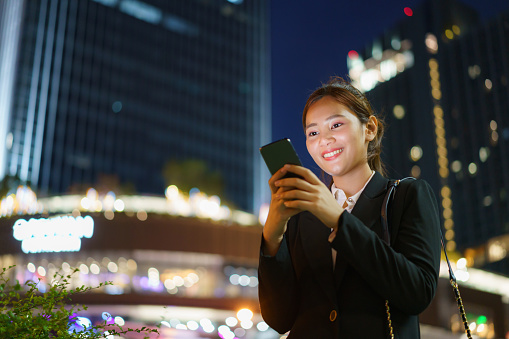 Asian executive working woman using a mobile phone in the street with office buildings in the background at night in Bangkok, Thailand.