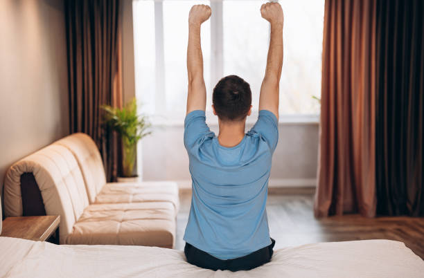 istock Good morning! Young man waking up in bed and stretching his arms, back view, copy space 1299193126
