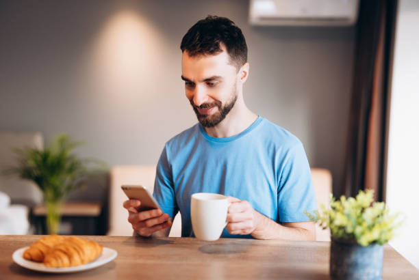 istock Daily life of young man in kitchen during breakfast using smartphone holding a cup of coffee. Attractive male working from home, telecommuting using remote internet job online communication on. 1299193152