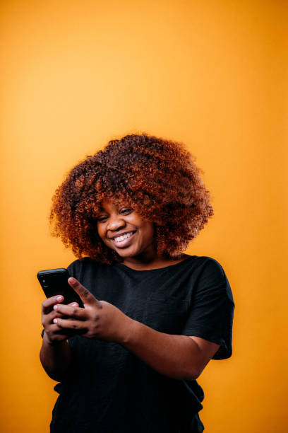 istock Studio Portrait of a Confident, Beautiful Young African American Woman Using a Mobile Handheld Smartphone to Send a Text Message, Online Bank, Online Shop, or Use Social Media in Front of a Happy Mustard Yellow-Colored Background 1299197788