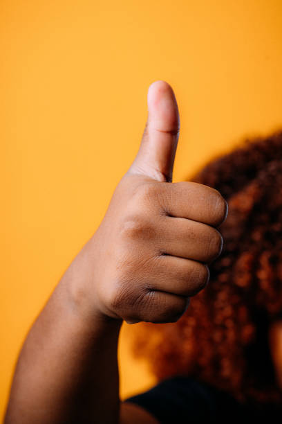 junge afroamerikanische frau rechte hand macht ein "daumen hoch" zeichen der zustimmung oder obszöne geste vor einem glücklichen gelben hintergrund - thumbs up african descent teenager women stock-fotos und bilder