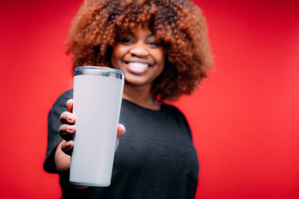 Studio Portrait of a Young African American Woman Holding or Serving a Generic Plain White Stainless Steel Coffee Mug in front of a Red Background - Focused on Foreground stock photo