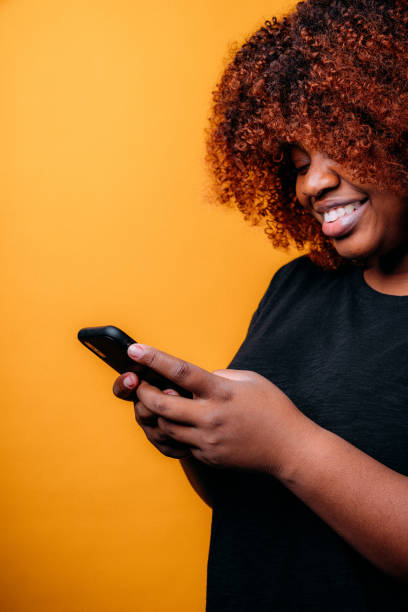 istock Studio Portrait of a Confident, Beautiful Young African American Woman Using a Mobile Handheld Smartphone to Send a Text Message, Online Bank, Online Shop, or Use Social Media in Front of a Happy Mustard Yellow-Colored Background 1299201142