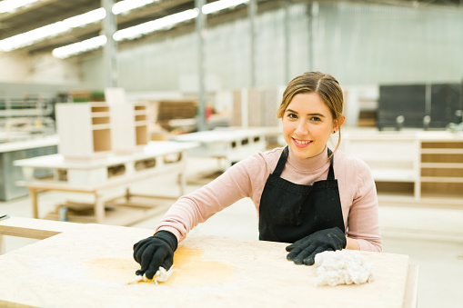 Portrait of a hard-working female carpenter smiling and staining wood with wood oil stain in a big spacious workshop