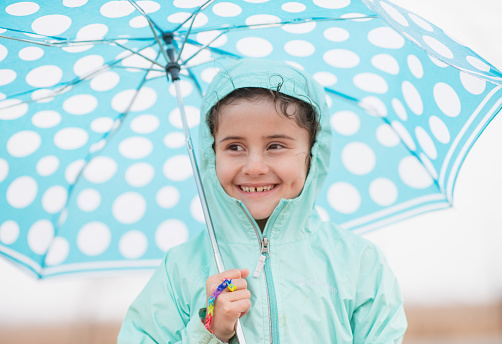 A super cute girl around 7 years of age is wearing a turquoise coloured rain coat and holding a blue polka dot umbrella. She is smiling.