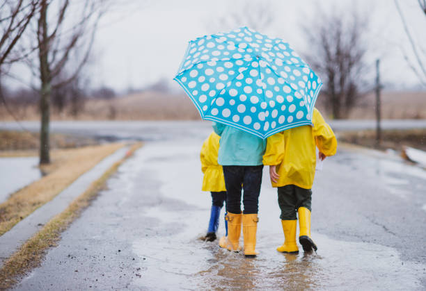 Siblings walking outdoors on a rainy day Back view of three siblings walking in the rain wearing raincoats and sharing a blue polka dot umbrella. raincoat stock pictures, royalty-free photos & images