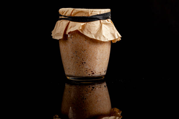 A glass cup of sourdough starter culture for homemade bread making. A glass cup of sourdough starter culture for homemade bread making. The cup is covered with brown filter paper and tightened with elastic band. Active culture creates bubbles. On reflective surface. yeast starter stock pictures, royalty-free photos & images