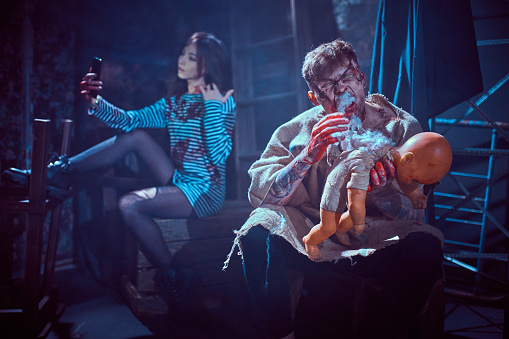A young man is dressed and made up like a zombie. A young beautiful woman ( girlfriend of a young man) with long dark hair is dressed striped frock. The zombie man is sitting and holding a baby doll. He is hungry and going to eat the doll. The young woman is sitting behind the zombie man. She is looking at mobile phone making selfie. Studio shooting in dark with light haze