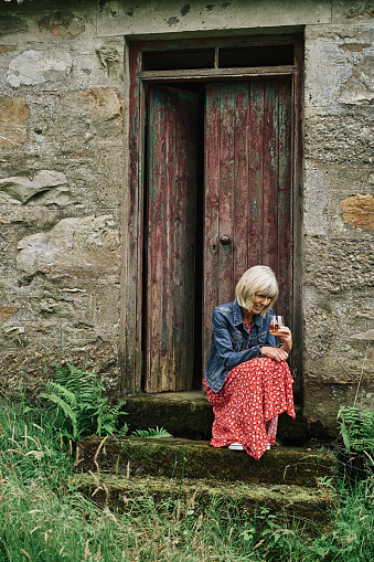 Mature woman relaxing with a glass of whisky outside a ruined bothy in Highland Scotland.