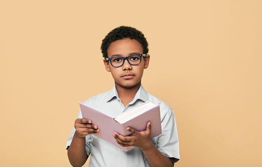 Serious African American little boy wearing eyeglasses holds book looking pensive at camera, on beige background