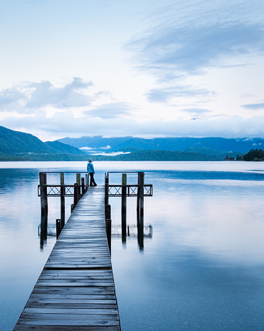 Lonely woman standing at the end of Lake Te Anau jetty, looking at the Murichison mountains. Vertical format.