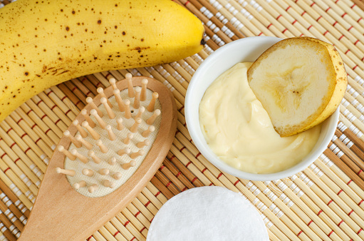 Yellow facial mask (banana face cream, shea butter hair mask, body butter) in the small white bowl and wooden hairbrush. Natural skin and hair concept. Top view, copy space.
