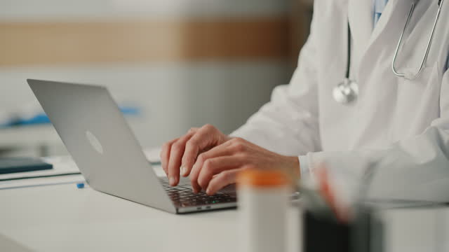 Experienced Middle Aged Male Doctor Wearing White Coat Working on Laptop Computer at His Office. Senior Medical Health Care Professional Working with Test Results, Patient Treatment Planning.