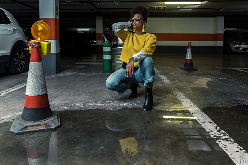 Full body of modern confident African American fashionable female model in trendy yellow shirt and accessories hunkering near puddle on underground parking lot