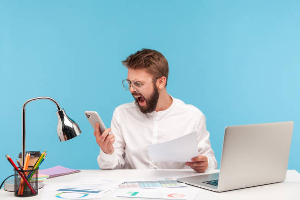 istock Angry nervous man office worker in white shirt screaming looking at smartphone display, skipping important event through lot of paper work 1299249611