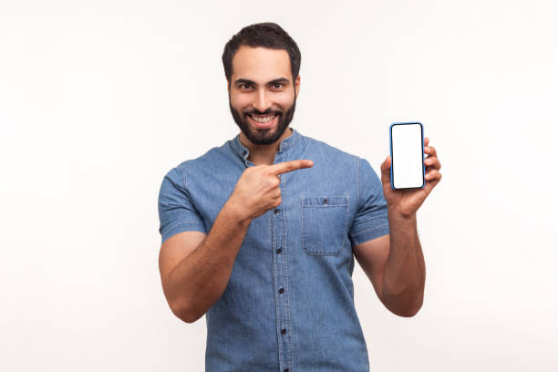 istock Happy cheerful beaded man in blue shirt pointing finger at smartphone with empty white screen and looking at camera with toothy smile, freespace for adv 1299249642
