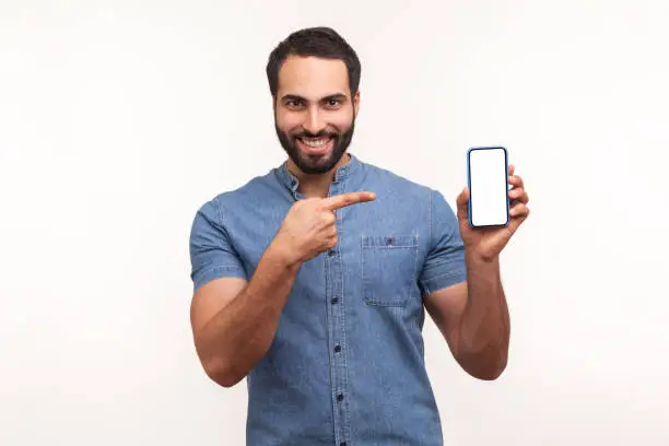 Happy cheerful beaded man in blue shirt pointing finger at smartphone with empty screen looking at camera with toothy smile, freespace for adv. Indoor studio shot isolated on white background