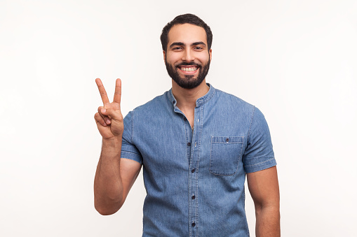 Happy satisfied man showing v sign symbol of peace with fingers, looking at camera with toothy smile, celebrating his successful victory. Indoor studio shot isolated on white background