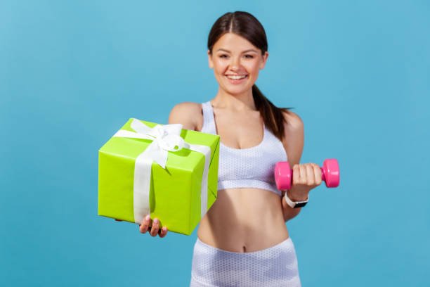 happy athletic woman in white sportswear holding green gift box and pink dumbbell in hand, preparing sports equipment as presents on holidays - muscular build bicep women female imagens e fotografias de stock