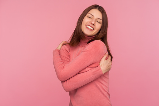 Positive narcissistic woman with brown hair tightly embracing herself standing with closed eyes, calming and supporting herself. Indoor studio shot isolated on pink background