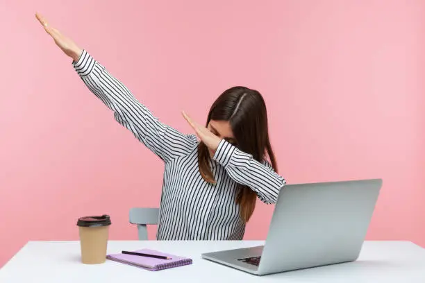 Photo of Extremely excited happy woman office worker showing dab dance gesture, performing internet meme of success, sitting at workplace with laptop.