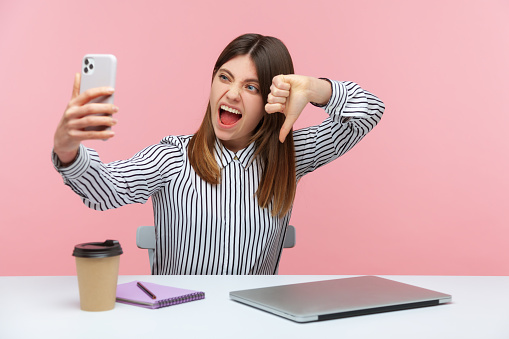 Excited crazy woman blogger in striped shirt showing thumbs down dislike gesture looking at smartphone camera, criticizing and hating. Indoor studio shot isolated on pink background