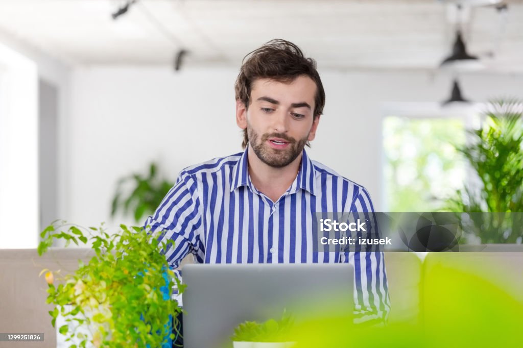 Bearded man using laptop in a green office Mid adult men wearing striped shirt sitting in the creative workplace, using laptop during video conference. Front View Stock Photo