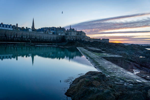 This photo was taken during the sunset in winters in Saint-Malo.  We can see the reflection of the ramparts in the water.  There is also a seagull which flies over intramural. The sunset gives contrast to the photo.  On the right side we can see the sea.  In the center we can also see the church and the roofs of Saint-Malo.  Finally, on the left side you can see the breakwaters, an emblem of Saint-Malo.