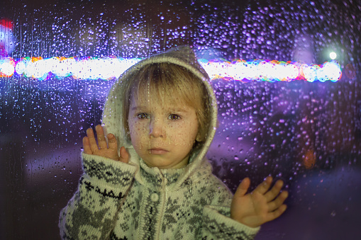 Little child, toddler boy, standing behind window with drops outdoors in the snow, christmas time