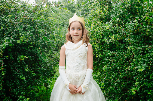 Outdoor portrait of elementary age girl wearing princess dress and paper crown standing in wet garden looking at camera