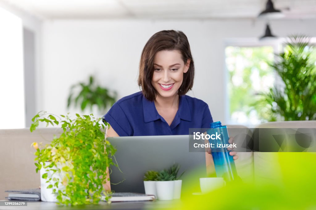 Smiling woman working on laptop at home Mid adult women sitting on sofa in the creative eco-friendly green workplace and using laptop during video call, holding water bottle. Laptop Stock Photo