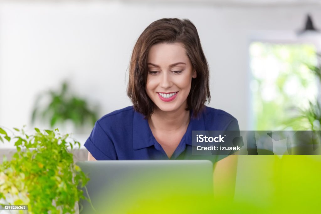 Smiling woman working on laptop at home Mid adult women sitting on sofa in the creative eco-friendly green workplace and using laptop during video call. Laptop Stock Photo