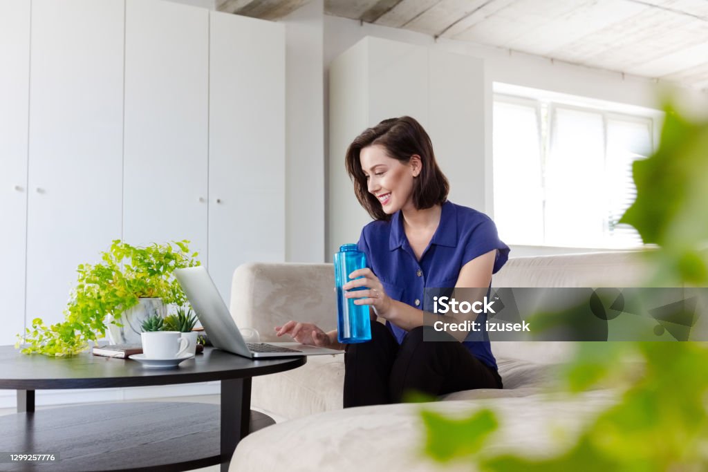 Woman working in the eco-friendly green office Mid adult women sitting on sofa in the creative workplace and using laptop, holding water bottle in hand. Office Stock Photo
