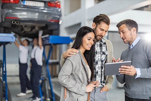 Mid adult salesman showing buying plans to young couple on digital tablet in auto repair shop.