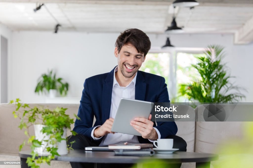 Businessman during video call Cheerful mid adult man wearing navy blue jacket sitting on sofa in the creative workplace and using digital tablet during video call. Environmental Conservation Stock Photo