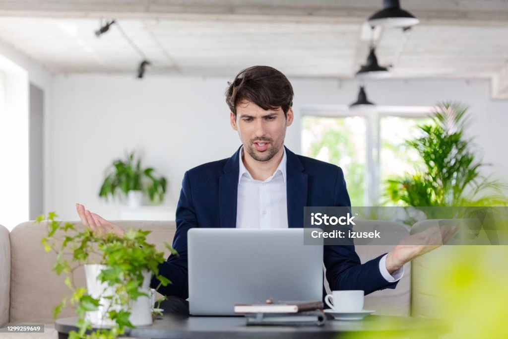 Businessman during video conference Displeased mid adult man wearing navy blue jacket sitting on sofa in the creative workplace and using laptop during video call. Displeased Stock Photo