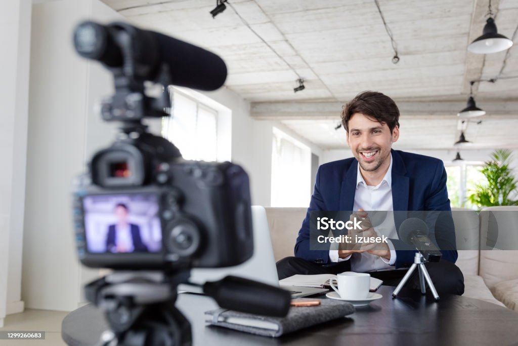 Businessman making a video blog in the office Mid adult businessmen wearing navy blue suit making a video blog. Vlogger recording content on digital camera in the creative office. Elegance Stock Photo