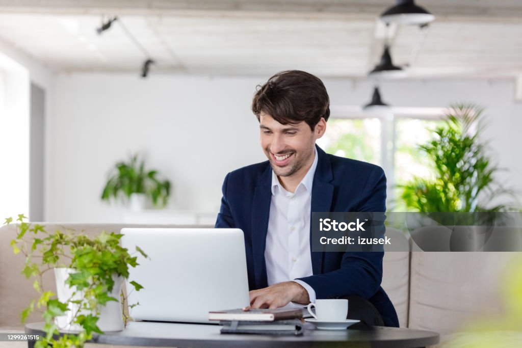 Businessman working on laptop in the green office Cheerful mid adult man wearing navy blue jacket sitting on sofa in the creative workplace and using laptop. Blue Stock Photo