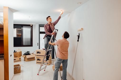 Young man on a stairway changing a light bulb while his friend helps to hold the ladder. Gay couple moved in a new home and is doing renovations. Apartment with cardboard boxes.
