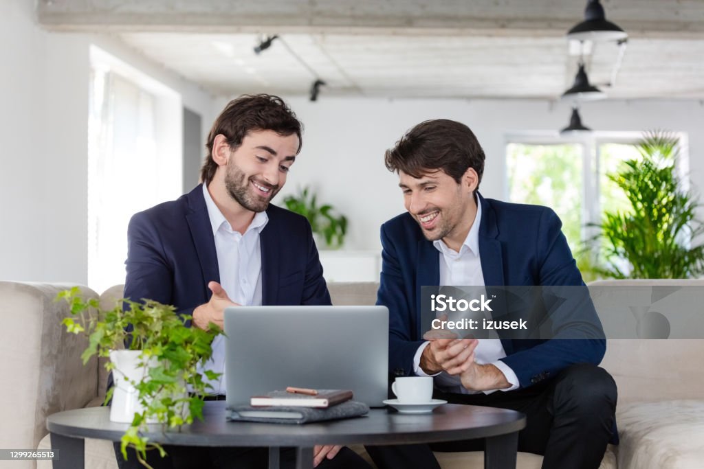 Shareholders discussing in an office Smiling mid adult businessmen wearing navy blue jackets and white shirts sitting on sofa in the green office and discussing over laptop. 30-39 Years Stock Photo