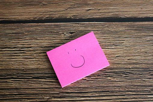 Close-up studio shot of a small pink adhesive notepad with a smiley face drawn on it on top of a wood table with copy space.