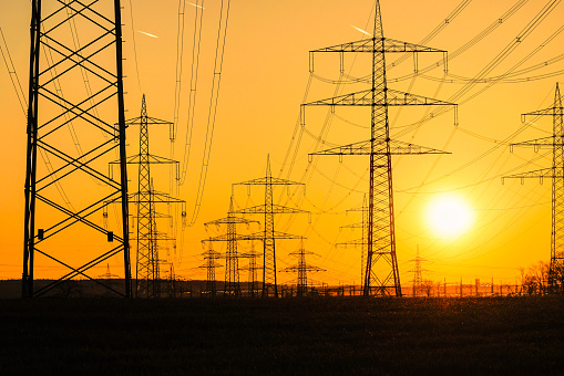 Electricity poles and electric power transmission lines against vibrant orange sky at sunset on a hot day with flickering air. High Voltage towers provide power supply over a long distance.