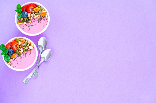 Healthy food backgrounds: overhead view of two yogurt bowls with blueberries, almonds, pumpkin seeds and chia seeds shot on purple background. Two metal spoons are beside the yogurt bowls. The composition is at the left of an horizontal frame leaving useful copy space for text and/or logo at the right. High resolution 42Mp studio digital capture taken with Sony A7rII and Sony FE 90mm f2.8 macro G OSS lens
