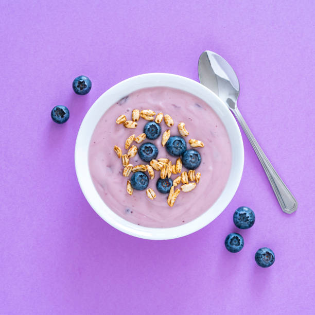 Healthy food backgrounds: overhead view of a white bowl filled wit yogurt, blueberries and cereal shot on purple background. A metal spoon is beside the bowl. Predominant colors are purple and white. High resolution 28Mp studio digital capture taken with Sony A7rII and Sony FE 90mm f2.8 macro G OSS lens