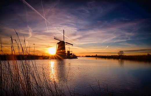 Dutch windmills during sunrise. The sun is already a bit higher in the sky and the sky is partially yellow and blue. There are quite some clouds. In the foreground is grass and on the other side of the canal are the windmills. The sky and the windmills are reflected in the water. Located in Kinderdijk, the Netherlands.
