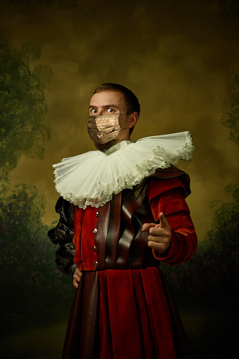 Portrait of medieval young man in vintage clothing and golden face mask standing on dark background. Male model as a royal person protected from covid. Concept of comparison of eras, modern, fashion.
