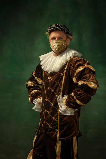 Portrait of medieval young man in vintage clothing, golden face mask posing confident, serious on dark background. Royal person protected from covid. Concept of comparison of eras, modern, fashion.